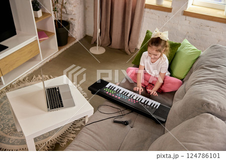 Little girl practicing keyboard on couch while watching online music class on laptop. Child studying at home, preschool preparation. 124786101