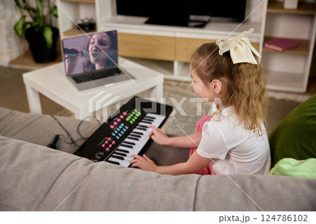 Girl watching online vocal training session while playing keyboard on sofa with focus and joy. Music lesson. Girl watching online vocal training session while playing keyboard on sofa with focus and joy. Music lesson. 124786102