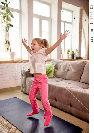 Excited girl jumping on mat during online physical class in bright cozy home setting. Online training. Excited girl jumping on mat during online physical class in bright cozy home setting. Online training. 124786103