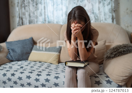 Woman reading bible, head bowed, praying while sitting on sofa with clasped hands 124787013