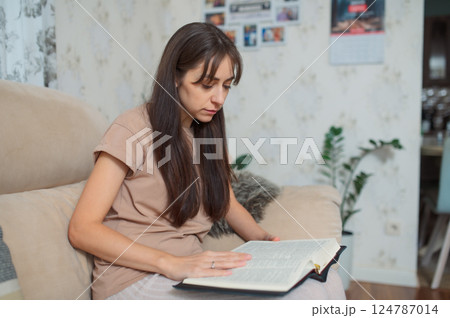 Millennial woman reading book bible while relaxing on comfortable sofa, immersed in quiet moment of solitude at home 124787014