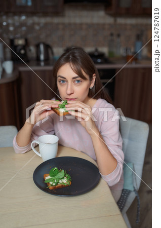 Young woman eating avocado toast healthy food, drinking herbal tea near bright kitchen window 124787019
