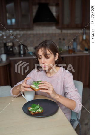 Young woman eating avocado toast healthy food, drinking herbal tea near bright kitchen window 124787020