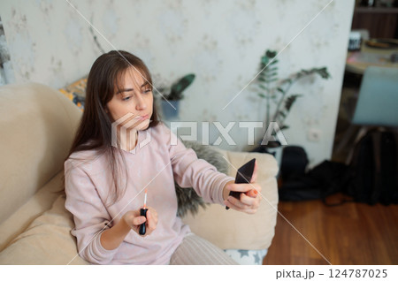 Young woman applying lipstick while looking at hand mirror while relaxing on living room sofa with soft background lighting - morning routine Young woman applying lipstick while looking at hand mirror while relaxing on living room sofa with soft background lighting - morning routine 124787025