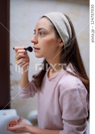 Woman 20s in bathrobe applying makeup, brushing cosmetics while standing before bathroom mirror morning routine 124787030