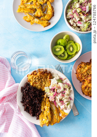 A healthy breakfast featuring brown rice with baked white fish, zucchini-carrot pancakes, Chinese cabbage salad with radish, and a refreshing kiwi dessert for a balanced, nutritious start to the day 124788766