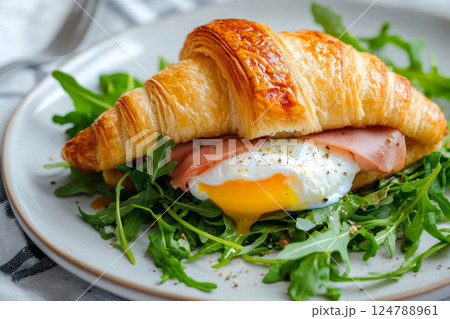 A golden, flaky croissant filled with a perfectly poached egg, ham, and fresh arugula, served on a rustic wooden board. The yolk is slightly runny, creating a rich and indulgent breakfast A golden, flaky croissant filled with a perfectly poached egg, ham, and fresh arugula, served on a rustic wooden board. The yolk is slightly runny, creating a rich and indulgent breakfast 124788961