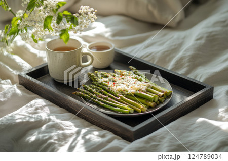 A dark modern wooden tray with roasted asparagus, Parmesan cheese, and chili flakes, accompanied by a cup of fragrant hot tea with mint leaves. Set on a white linen bed with soft sunlight filling 124789034