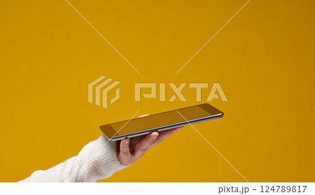 Hand wearing a white sweater holds a silver tablet against a solid yellow background. The tablet screen is blank and facing upwards, copy space 124789817