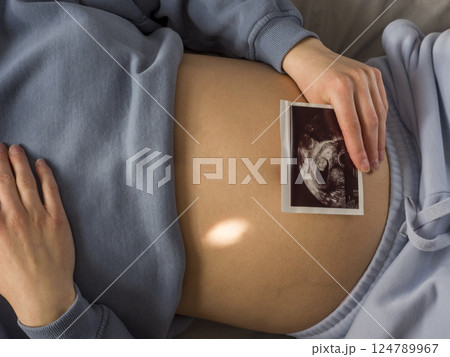 Pregnant woman tenderly holding ultrasound photo near belly, softly caressing abdomen while bonding with expecting baby in peaceful bedroom setting Pregnant woman tenderly holding ultrasound photo near belly, softly caressing abdomen while bonding with expecting baby in peaceful bedroom setting 124789967