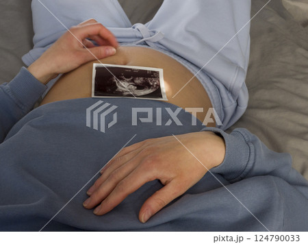 Expectant mother resting in blue attire, gently resting ultrasound image on pregnant belly, radiating maternal warmth and anticipation of new life Expectant mother resting in blue attire, gently resting ultrasound image on pregnant belly, radiating maternal warmth and anticipation of new life 124790033