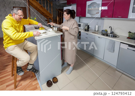 Wife serves food at kitchen island while husband sits on bar stool nearby, pet bowls sit on floor in spacious kitchen with wooden accents. 124793412