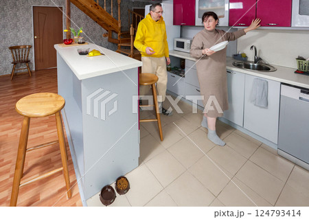 Wife holds plate near sink with red cabinets, husband stands at island with gray cabinets, stairs and pet bowls visible in brightly lit kitchen. 124793414
