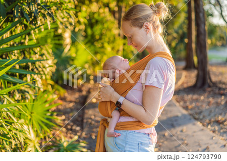 A loving mother walks through a peaceful park, carrying her baby securely in a sling. A heartwarming moment of maternal bonding, babywearing, and the joy of motherhood in nature 124793790