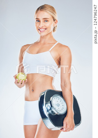Scale, smile and portrait of a woman with an apple to lose weight against a grey studio background. Health, nutrition and happy young model with fruit and scales for training motivation and wellness Scale, smile and portrait of a woman with an apple to lose weight against a grey studio background. Health, nutrition and happy young model with fruit and scales for training motivation and wellness 124796247