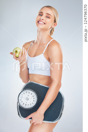 Health, lose weight and woman with an apple and scale for body goal, motivation and nutrition against a grey studio background. Food, happy and portrait of athlete model with smile for fruit and diet 124796370