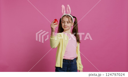 Enthusiastic small child spinning around in front of camera, presenting her red painted egg for easter sunday festivity. Cheery toddler with bunny ears twirling and doing a pirouette. Camera B. Enthusiastic small child spinning around in front of camera, presenting her red painted egg for easter sunday festivity. Cheery toddler with bunny ears twirling and doing a pirouette. Camera B. 124797024