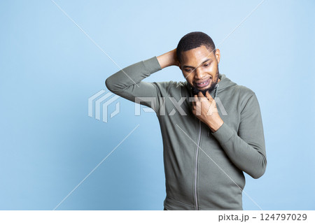 Clueless doubtful adult with a grey hoodie thinking about solutions and new ideas for innovation, being in his thoughts against blue background. African american man contemplating in studio. Clueless doubtful adult with a grey hoodie thinking about solutions and new ideas for innovation, being in his thoughts against blue background. African american man contemplating in studio. 124797029