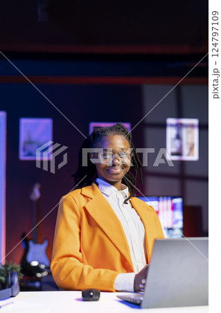 Portrait of jolly woman sitting on chair at home office desk in neon illuminated apartment late at night. Cheerful african american person in RGB lit living room enjoying leisure time Portrait of jolly woman sitting on chair at home office desk in neon illuminated apartment late at night. Cheerful african american person in RGB lit living room enjoying leisure time 124797109