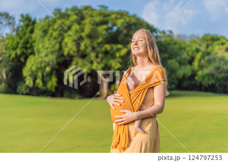 Mother holding her little baby in a yellow sling in the park. Warm and loving family moment. Babywearing, parenting, and mother-child bonding concept 124797253