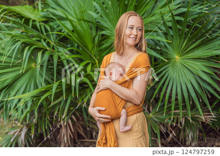 Mother holding her little baby in a yellow sling in the park. Warm and loving family moment. Babywearing, parenting, and mother-child bonding concept Mother holding her little baby in a yellow sling in the park. Warm and loving family moment. Babywearing, parenting, and mother-child bonding concept 124797259
