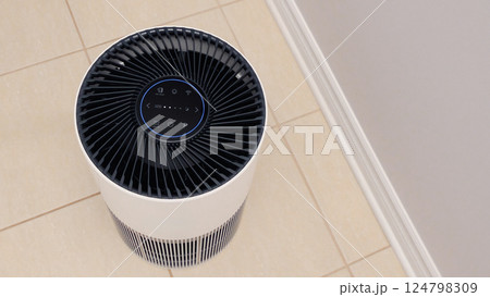 Tall round air purifier with a black top fan and a white body on a tile floor against the wall 124798309