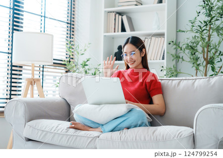 Happy young Asian woman wearing eyeglasses using laptop while seated on couch at home Happy young Asian woman wearing eyeglasses using laptop while seated on couch at home 124799254