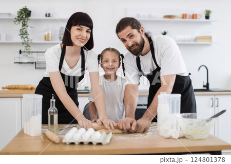 Caucasian family of parents and a young daughter cooking in modern kitchen, bonding, enjoying preparing and rolling dough together, showcasing teamwork, happiness, and quality time in a pleasant 124801378