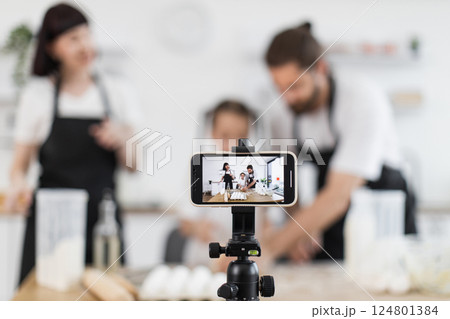 Caucasian family blogging their culinary activity, including dancing and dough kneading, in modern kitchen. Video captured on smartphone highlighting fun, creativity, teamwork. 124801384