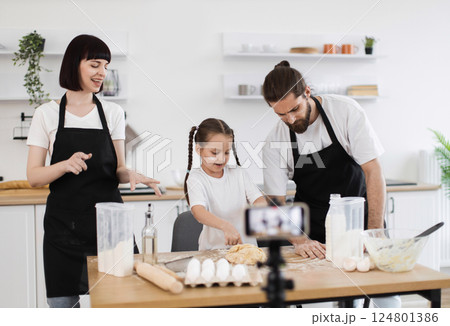 Caucasian family cooking together in kitchen, recording video while bonding over food preparation. Parents and child in aprons kneading dough, smiling and enjoying shared cooking experience. Caucasian family cooking together in kitchen, recording video while bonding over food preparation. Parents and child in aprons kneading dough, smiling and enjoying shared cooking experience. 124801386