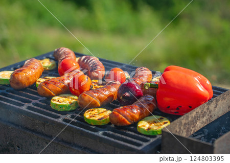 Delicious grilled sausages with vegetables on barbecue grill with smoke and flames in green grass Delicious grilled sausages with vegetables on barbecue grill with smoke and flames in green grass 124803395