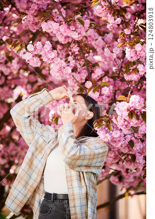 Woman allergic using medical eyes drops, suffering from seasonal allergy at spring in blossoming garden. Young woman treating eyes in front of blooming tree outdoors. Spring allergy concept. Woman allergic using medical eyes drops, suffering from seasonal allergy at spring in blossoming garden. Young woman treating eyes in front of blooming tree outdoors. Spring allergy concept. 124803918