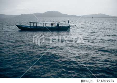A lone boat on a tranquil, gray sea with mountains in the background under an overcast sky during the monsoon season 124805513