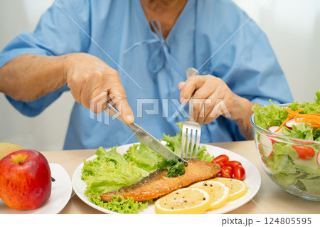 Asian senior woman patient eating Salmon steak breakfast with vegetable healthy food while sitting and hungry on bed in hospital. 124805595
