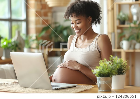 Young pregnant African American woman in front of laptop monitor at home. The young woman smiles as she connects with her doctor online, celebrating her pregnancy milestones. 124806651