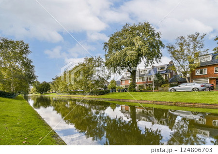 A serene riverside view featuring trees lining a calm waterway, complemented by charming nearby houses. The clear sky enhances the picturesque landscape. 124806703