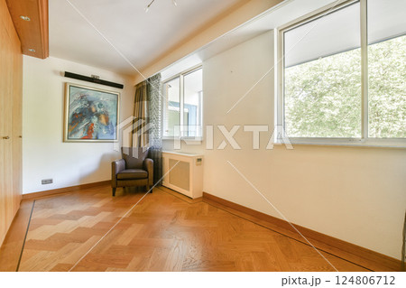 A well-lit modern interior room featuring a comfortable chair, artwork on the wall, and large windows offering natural light and a view of greenery outside. 124806712