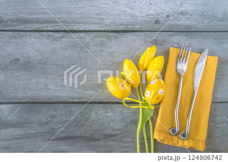 Place setting for easter dinner or lunch: bouquet of yellow tlips, easter eggs and fork and knife on a yellow napkin, wooden background; copy space 124806742