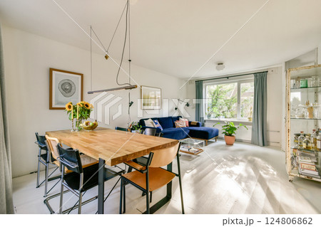 A stylish living space featuring a wooden dining table, blue sofa, and large windows bringing in natural light and greenery. A stylish living space featuring a wooden dining table, blue sofa, and large windows bringing in natural light and greenery. 124806862