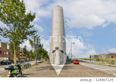 A striking modern building stands tall next to a serene waterfront, surrounded by trees and city life. Its unique design draws attention and complements the urban landscape. 124807129