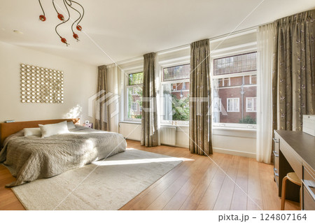 A stylish and inviting bedroom featuring large windows that allow natural light to flood the space, complemented by warm wooden flooring and decorative elements. A stylish and inviting bedroom featuring large windows that allow natural light to flood the space, complemented by warm wooden flooring and decorative elements. 124807164
