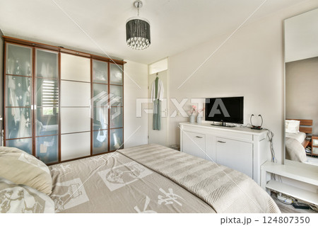 A stylish bedroom featuring a cozy bed, a sleek TV, wardrobe with sliding doors, and soft lighting. The room exudes a contemporary and relaxing atmosphere. 124807350