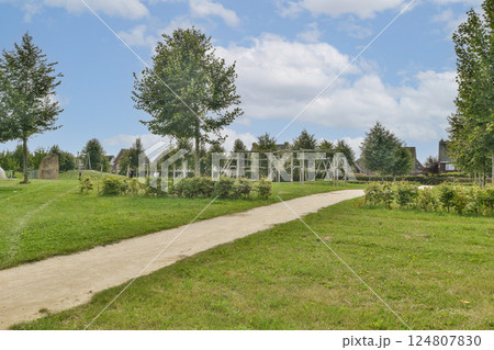 A peaceful park featuring a winding pathway, vibrant greenery, and playground equipment amidst tall trees under a cloudy sky, inviting visitors for relaxation and enjoyment. 124807830