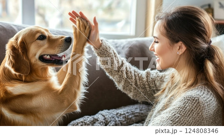 A woman and her golden retriever share a joyful high five while sitting together on a cozy couch at home 124808346