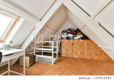 A modern attic workspace includes cozy decor, a desk, and organized storage. The room is brightened by natural light from a large window. A modern attic workspace includes cozy decor, a desk, and organized storage. The room is brightened by natural light from a large window. 124812448