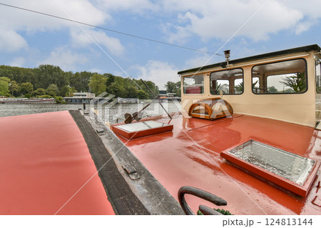 The image showcases a vibrant red boat docked by the water with lush greenery in the background. The sky is partly cloudy, adding to the serene atmosphere. 124813144