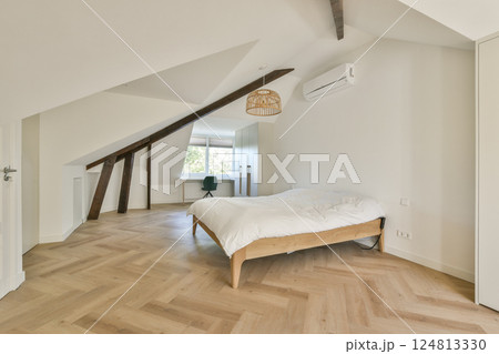 A stylish modern bedroom featuring a wooden floor, slanted ceiling, and a cozy bed. The space is well-lit with natural light from a nearby window. 124813330