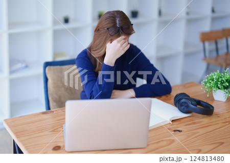 Stressed young woman overwhelmed by e-learning studies at desk with laptop, notebook, headphones, hands clasped to her face in frustration 124813480