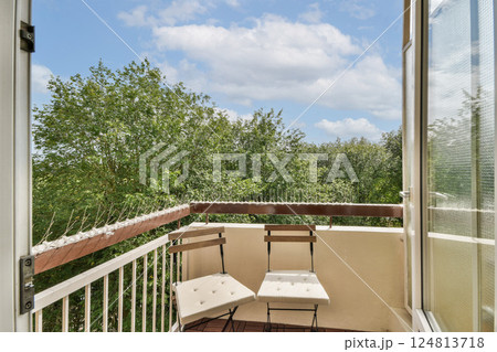 A peaceful balcony showcasing a light wooden railing and two chairs, overlooking lush green trees and a bright blue sky with clouds. 124813718