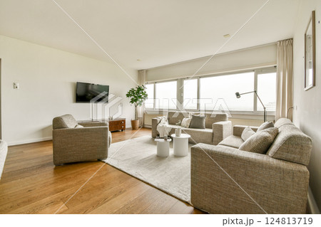 A spacious modern living room featuring light-colored walls, comfortable seating, a TV unit, and large windows that allow natural light to fill the space. 124813719